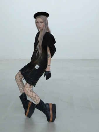 Model in black dress wearing fishnet tights and platform boots standing against white backdrop