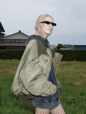 Model wearing dark sunglasses, olive green bomber jacket, and dark hoodie outdoors against an open landscape.