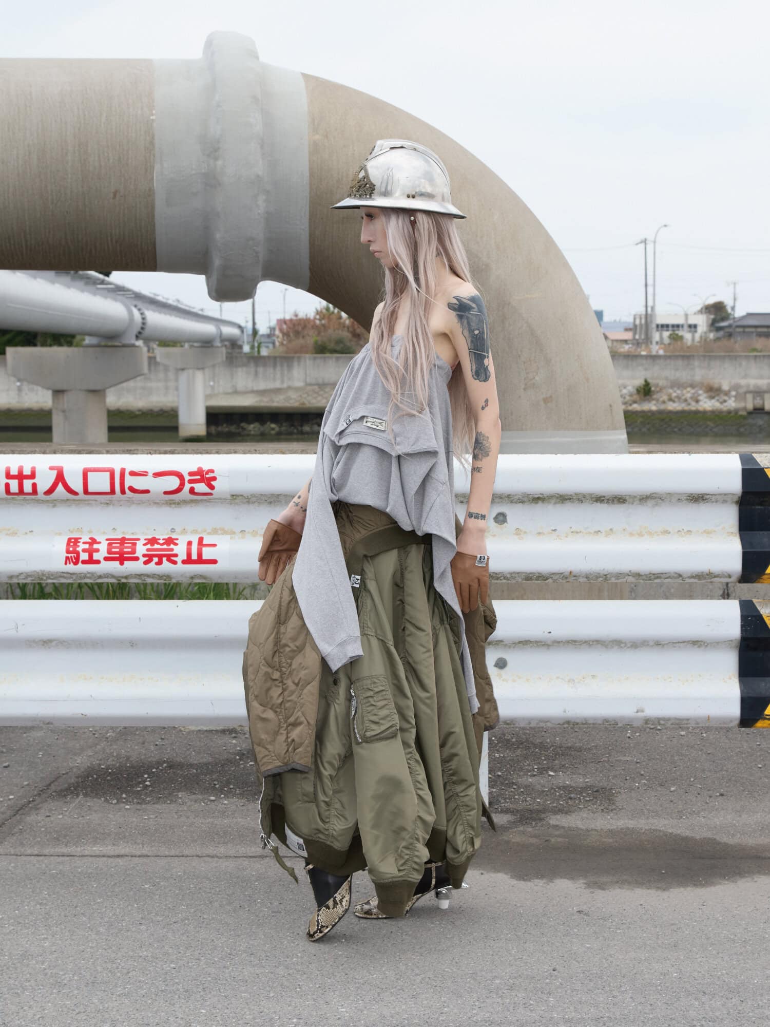 Model wearing a metallic fire helmet, grey draped top, and layered olive trousers against an outdoor industrial backdrop.