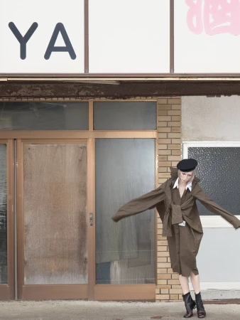 Model in oversized brown coat and black beret standing before a brick storefront.