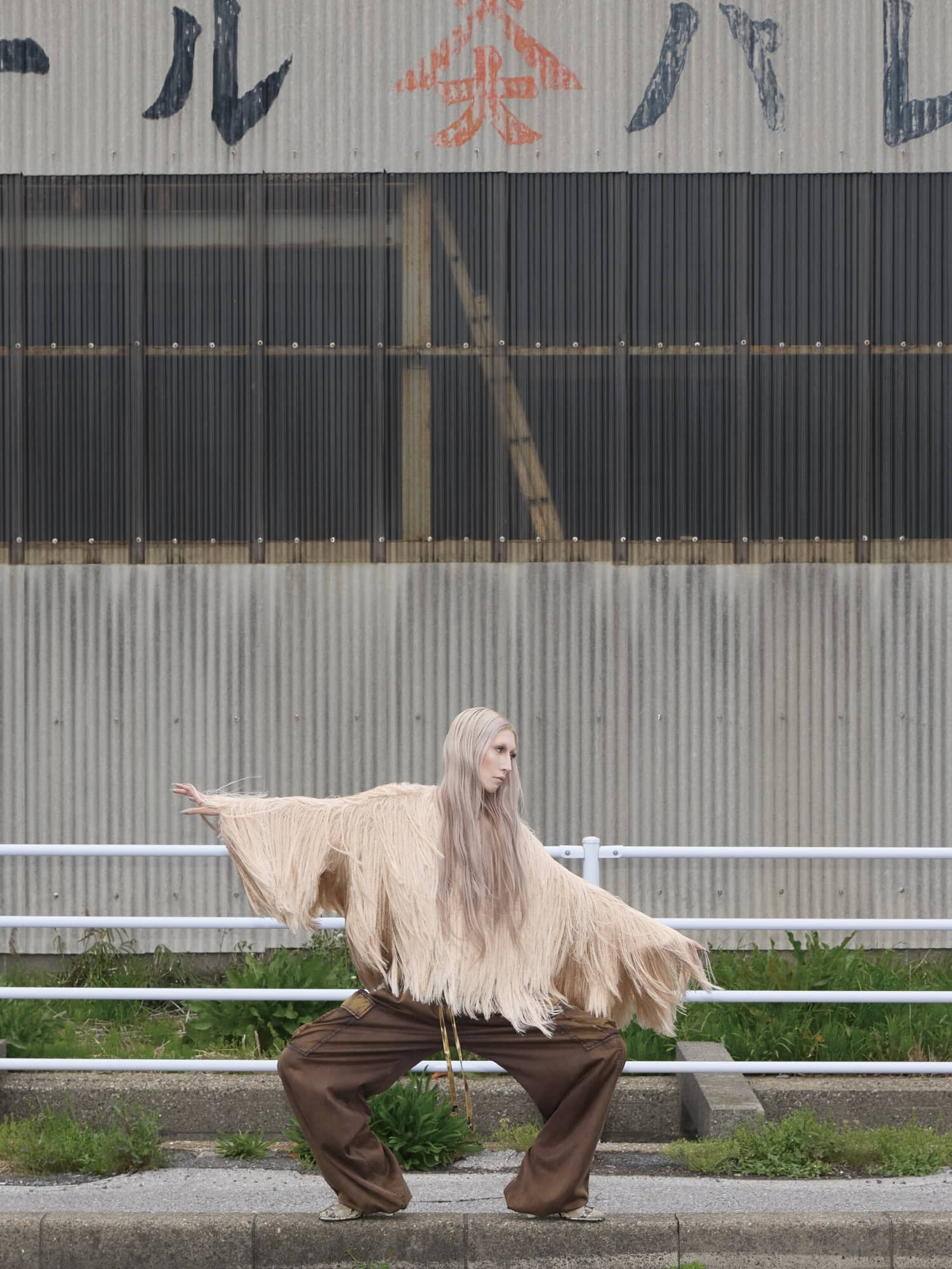 Model with long blonde hair in a fringed beige top and brown trousers posing before a corrugated metal wall