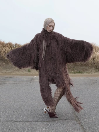 Model in long brown fringed textured garment and heels standing on asphalt against a grassy landscape