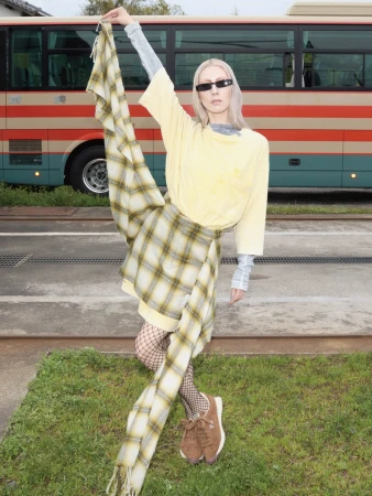 Model in layered yellow t-shirt and plaid skirt holding up a matching scarf in front of a bus