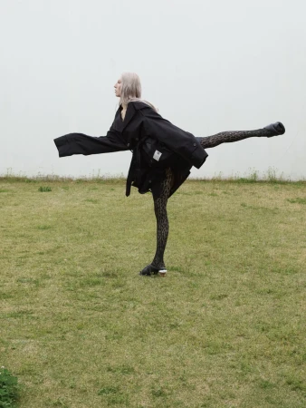 Model in a black coat and patterned tights balancing on one leg in a grassy field against an overcast sky