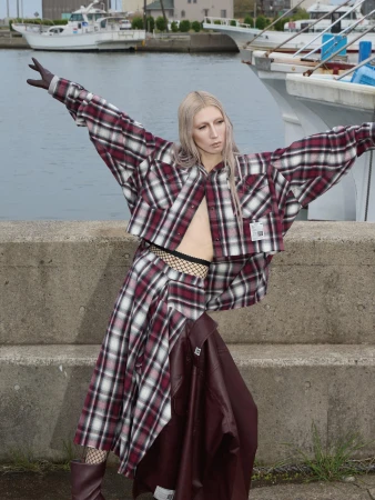 Model with long blonde hair wearing a plaid outfit and dark gloves standing against a concrete wall near docked boats.
