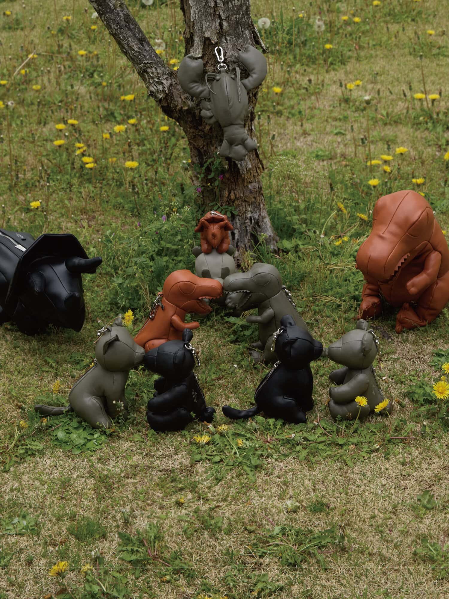 Collection of leather dinosaur and animal shaped bag charms arranged on a grassy patch around a tree trunk for Maison Mihara Yasuhiro.