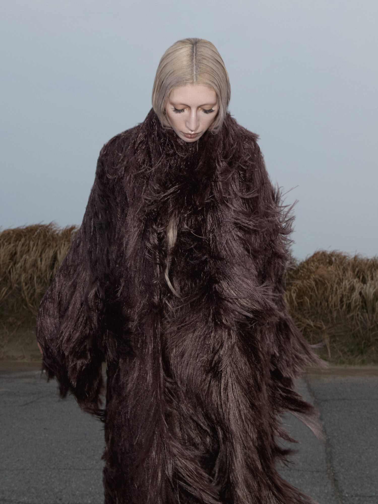 Model with blonde bob wearing a textured dark brown shaggy coat outdoors against an overcast sky