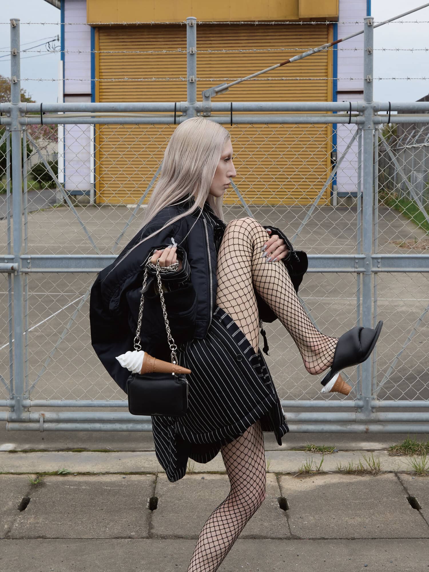 Model with long blonde hair wearing a black bomber jacket and fishnet tights holding a black handbag with an ice cream prop
