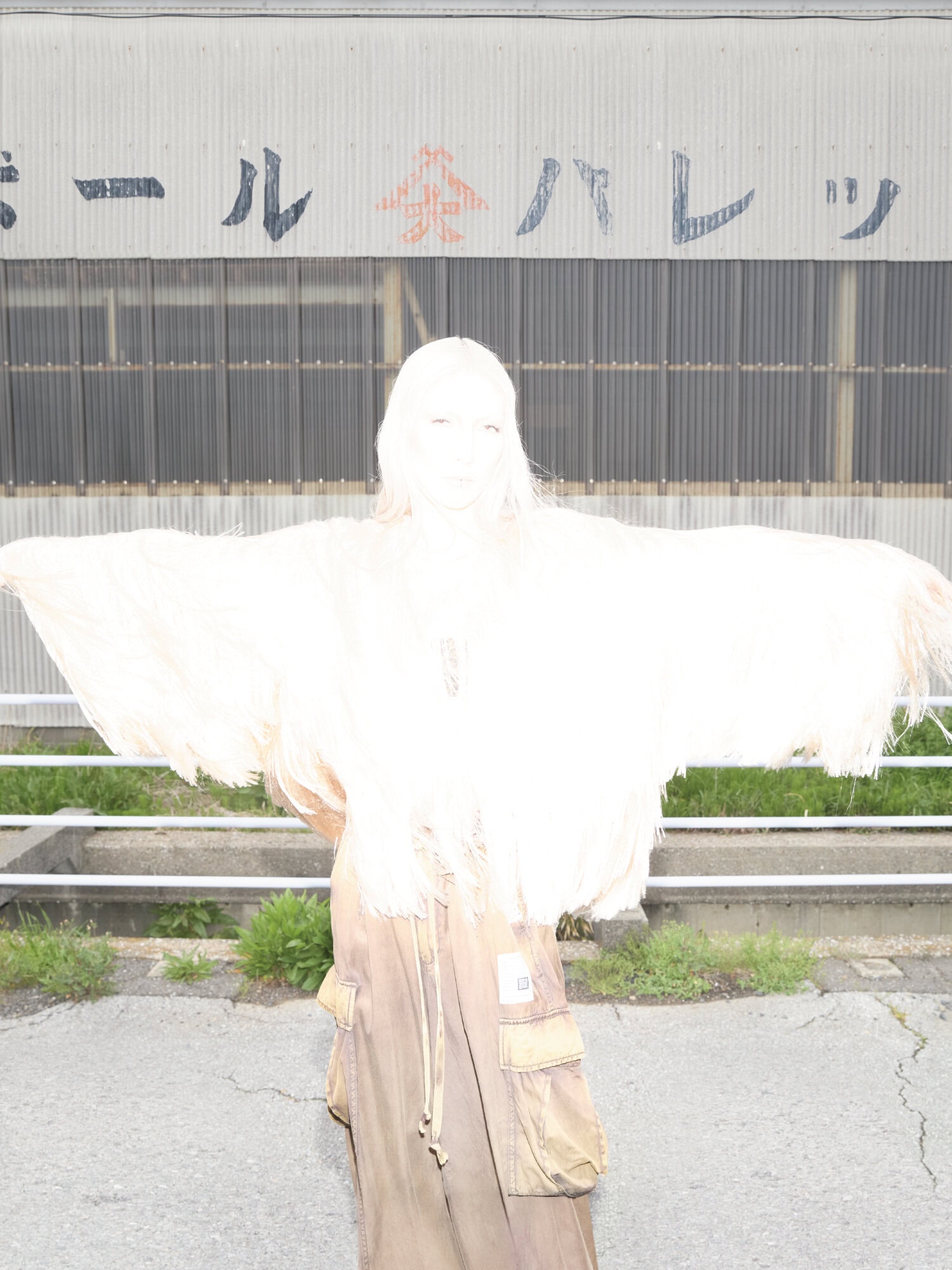 Model standing outdoors with arms outstretched wearing beige cargo pants and a light top against an industrial wall
