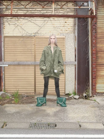 Model standing against an industrial building wearing an oversized olive green jacket and quilted green boots