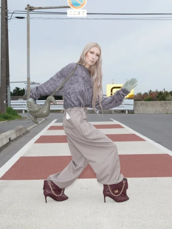 Model with long blonde hair in a grey knit sweater and wide taupe trousers posing on a striped crosswalk holding a duck-shaped bag