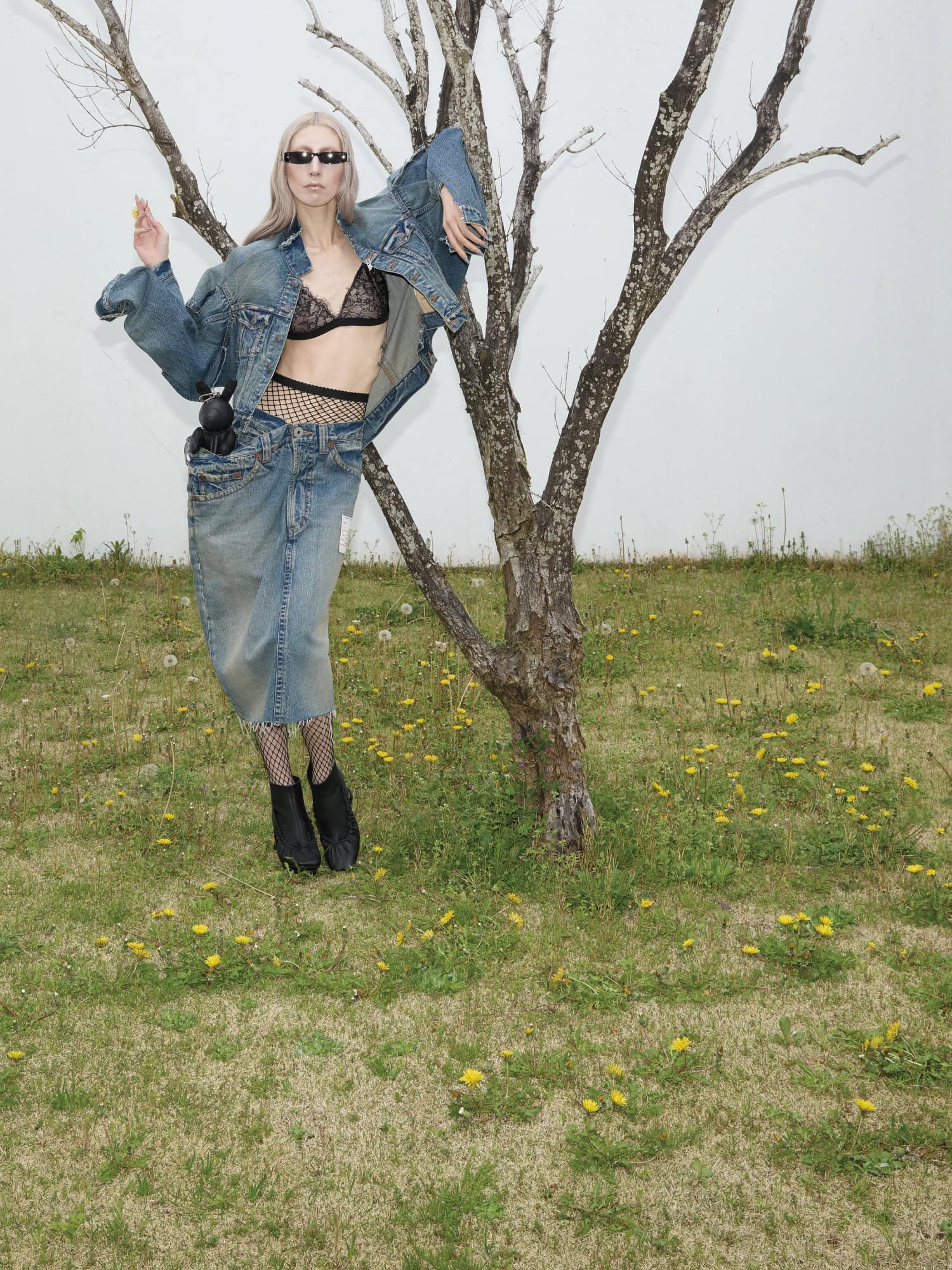 Model wearing oversized denim jacket and skirt leaning against a bare tree in a grassy meadow