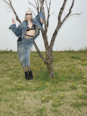 Model wearing oversized denim jacket and skirt leaning against a bare tree in a grassy meadow