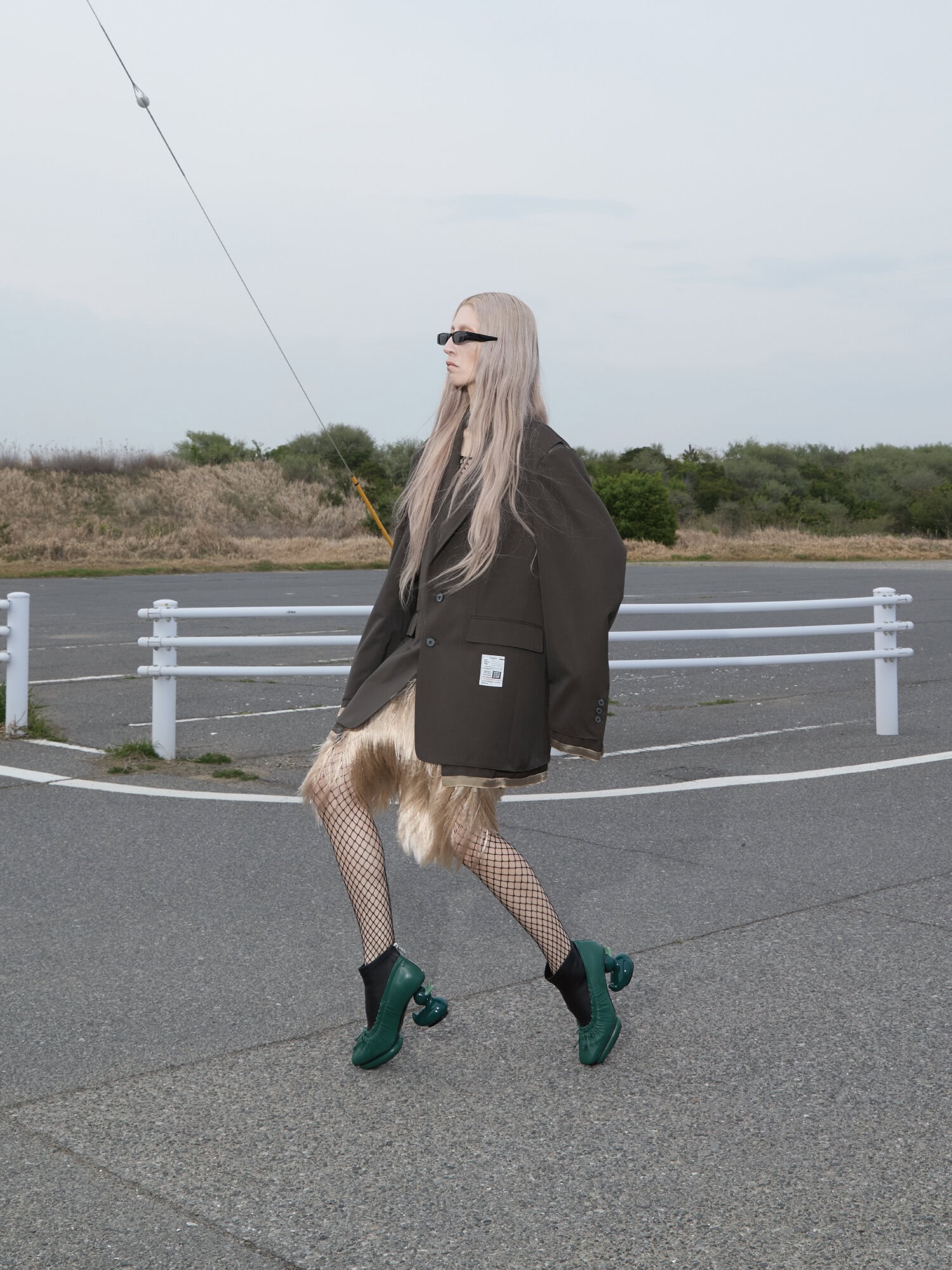 Model with long blonde hair wearing an oversized dark blazer and green sculptural heels on a paved tarmac surface.
