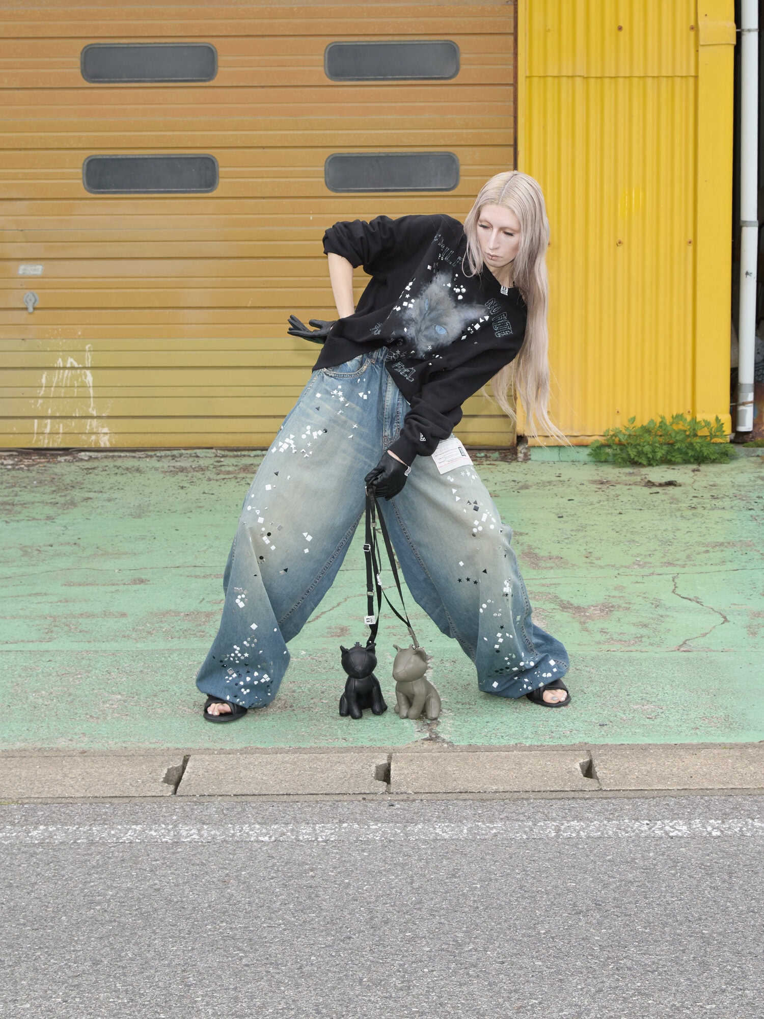 Model with long blonde hair in oversized black sweatshirt and paint-splattered baggy jeans posing outdoors with two toy dog figurines.