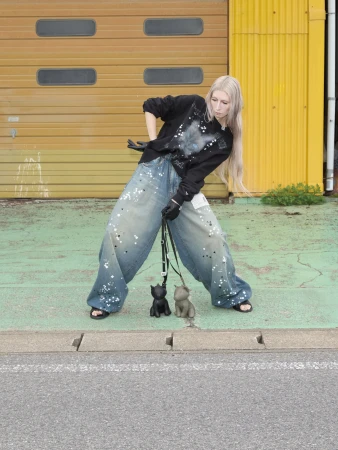 Model with long blonde hair in oversized black sweatshirt and paint-splattered baggy jeans posing outdoors with two toy dog figurines.
