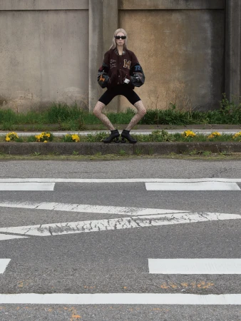 Model in sunglasses and varsity jacket squatting on a sidewalk curb next to a paved street for Maison Mihara Yasuhiro.