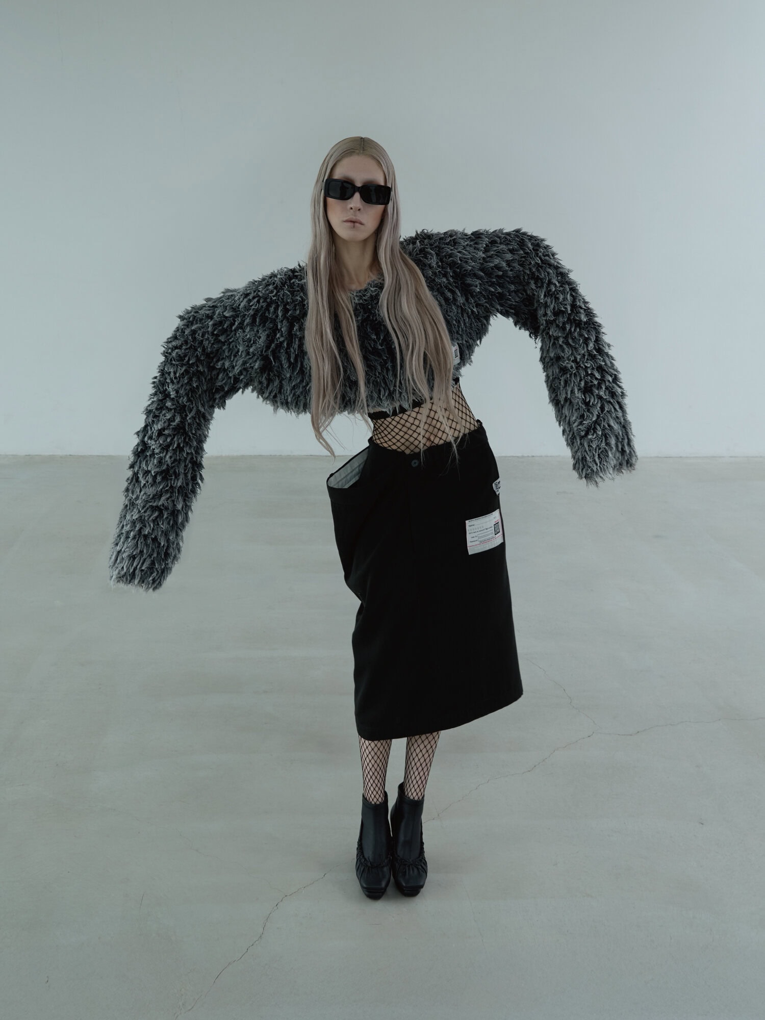 Model wearing dark sunglasses, fuzzy textured cropped sweater, black skirt, and fishnet stockings against white seamless backdrop