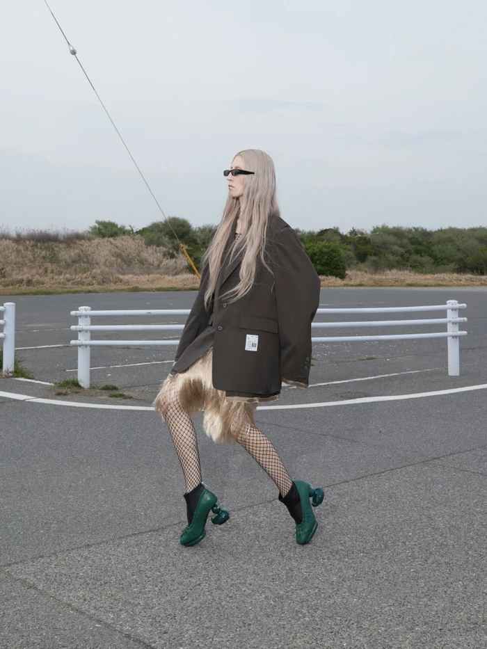 Model with long blonde hair wearing an oversized dark blazer and green sculptural heels on a paved tarmac surface.