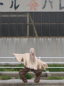 Model with long blonde hair in a fringed beige top and brown trousers posing before a corrugated metal wall