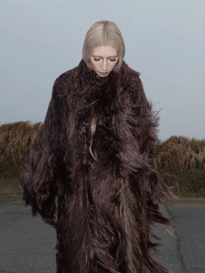 Model with blonde bob wearing a textured dark brown shaggy coat outdoors against an overcast sky