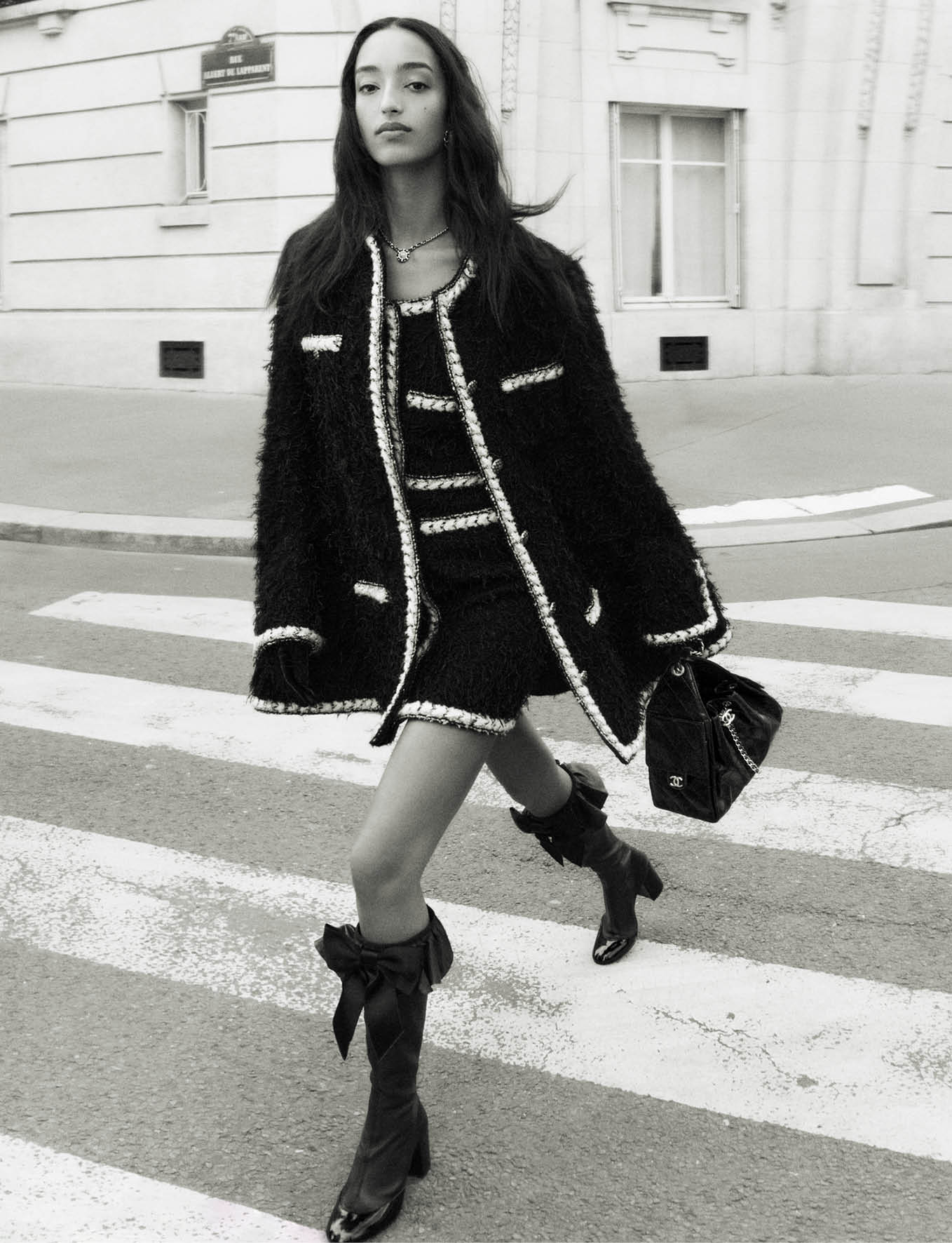 Model Mona Tougaard walking across a city street wearing a black tweed jacket and carrying a handbag for Chanel Fall 2025 campaign.