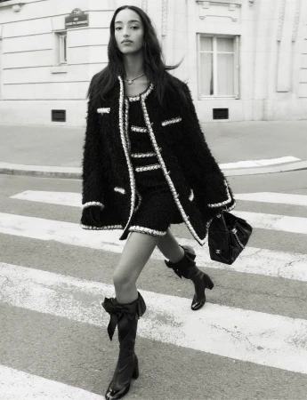 Model Mona Tougaard walking across a city street wearing a black tweed jacket and carrying a handbag for Chanel Fall 2025 campaign.