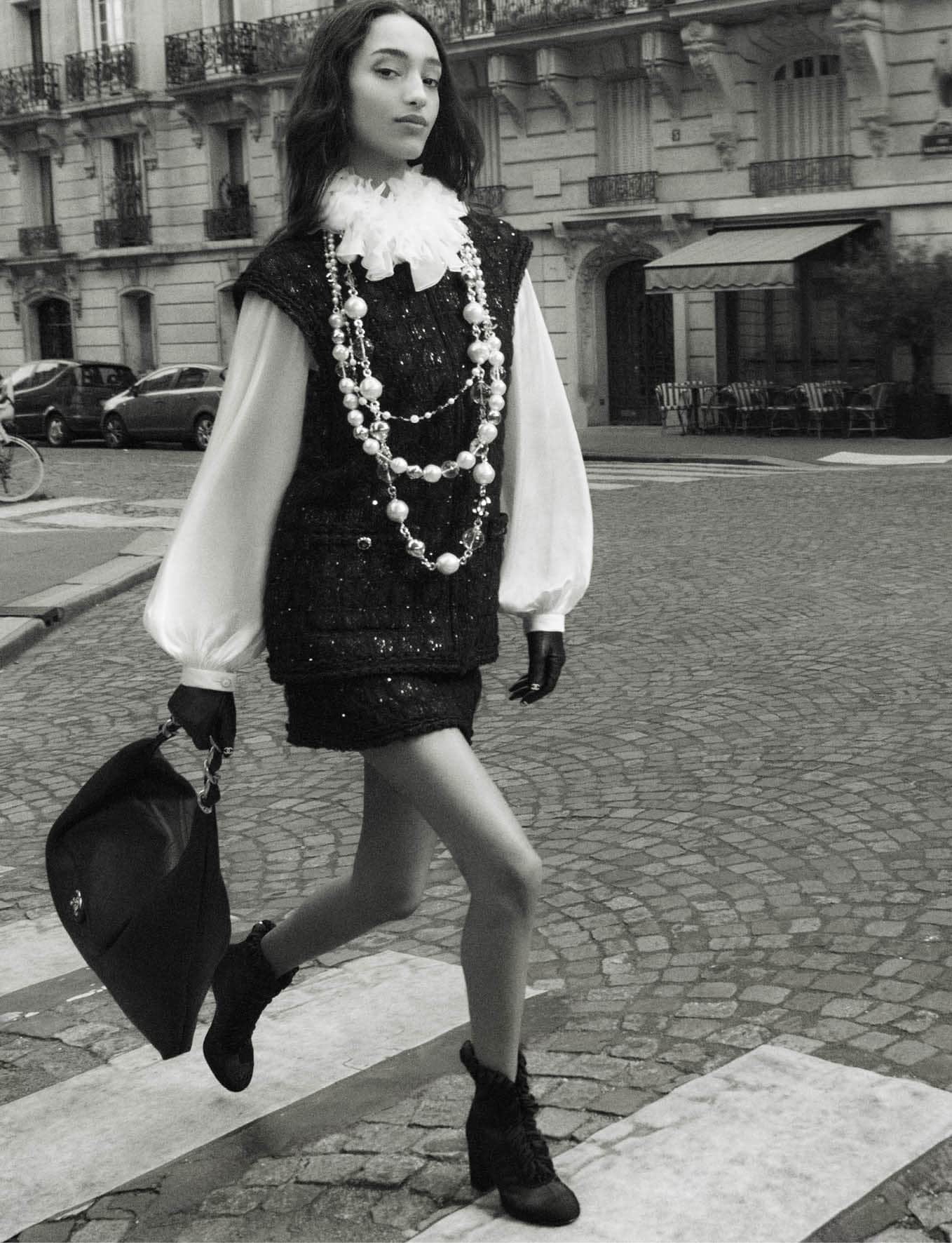 Mona Tougaard walking on a cobblestone street wearing a tweed vest and pearl necklaces for Chanel Fall 2025 ad campaign.