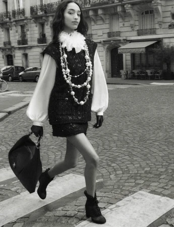 Mona Tougaard walking on a cobblestone street wearing a tweed vest and pearl necklaces for Chanel Fall 2025 ad campaign.