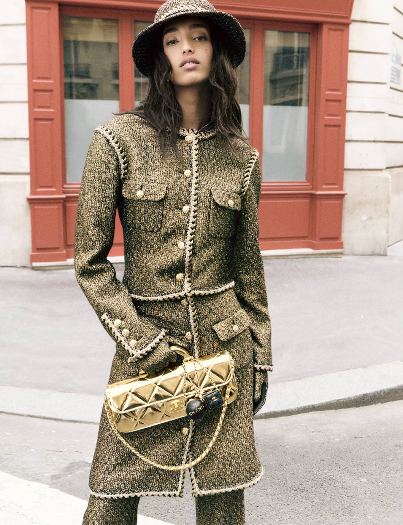 Model Mona Tougaard wearing a tweed suit and matching hat holding a gold quilted handbag on a city street.