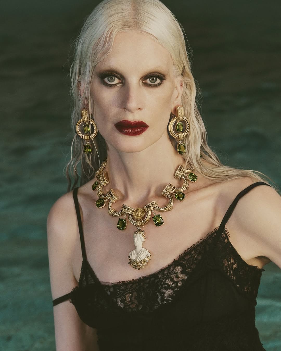 Kristen McMenamy wearing statement gold jewelry and a black lace dress against water for Dolce & Gabbana