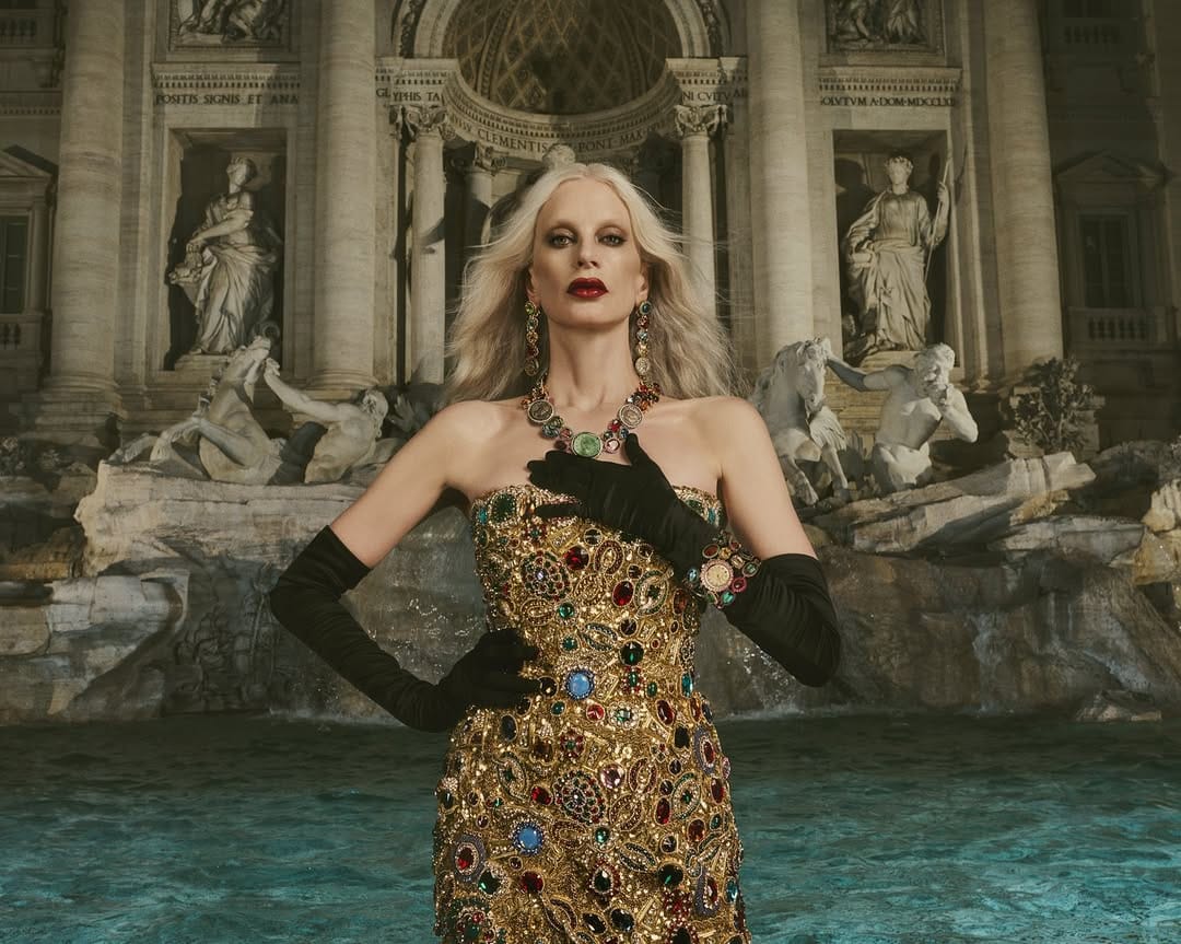 Kristen McMenamy wearing a jewel-embrusted gold dress and black opera gloves in front of a classical stone fountain.