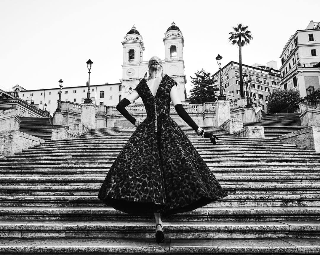 Model Kristen McMenamy in a patterned full skirt dress and opera gloves walking up grand stone outdoor stairs in Rome.