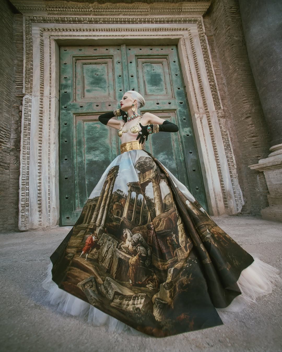Kristen McMenamy in a Dolce & Gabbana gown featuring a painted classical ruin skirt standing before large green doors