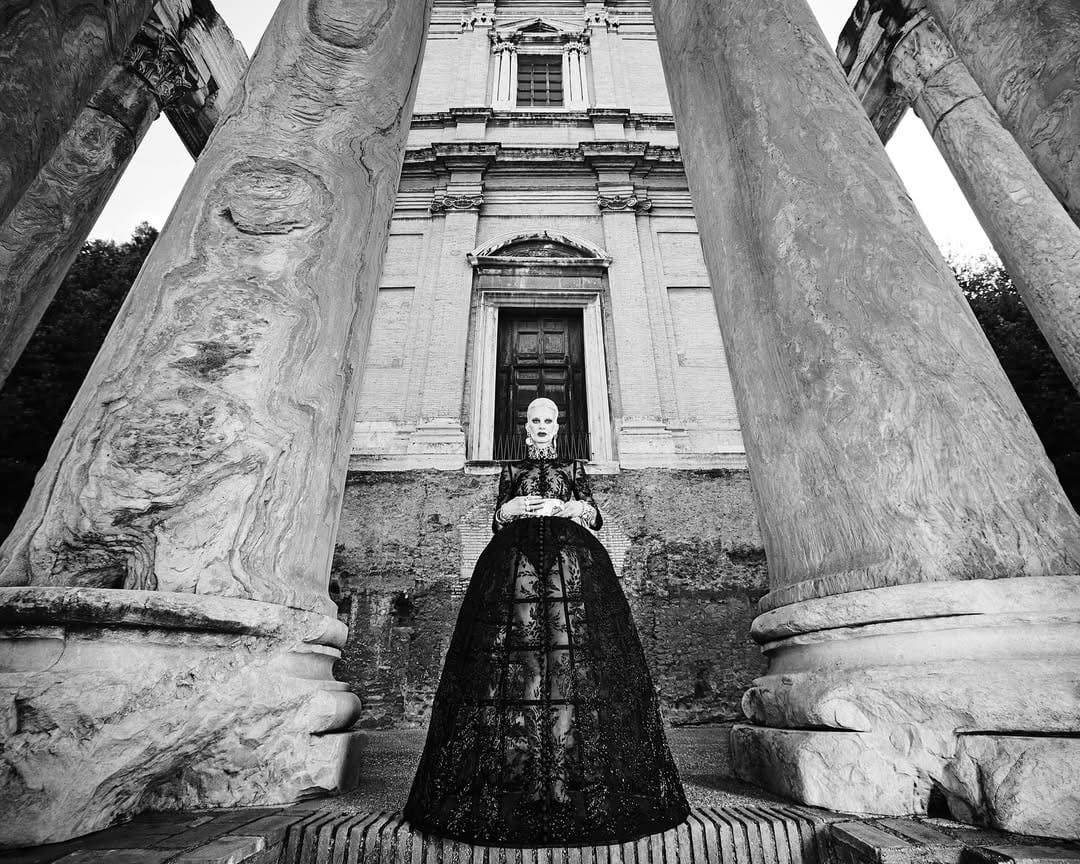 Model Kristen McMenamy in an ornate black lace gown standing between classical stone columns for Dolce & Gabbana Alta Moda