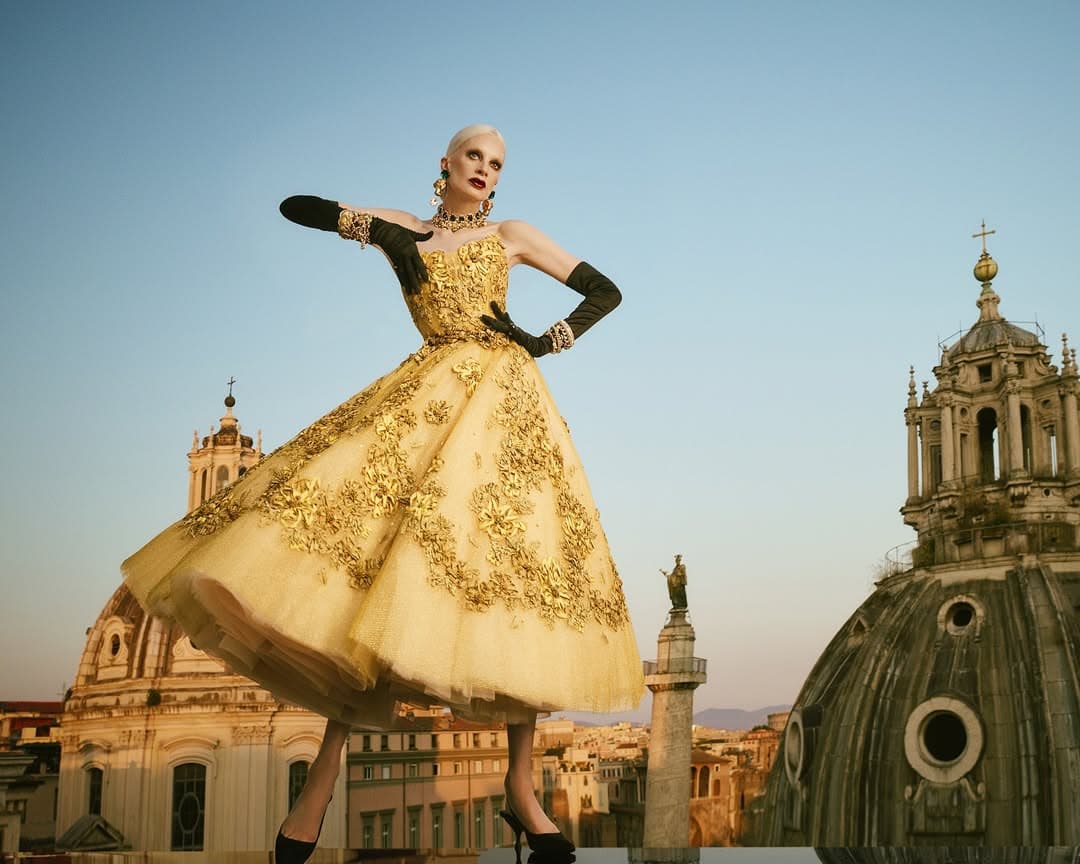 Kristen McMenamy in an ornate gold dress and black gloves on a rooftop with architectural domes under a clear sky.