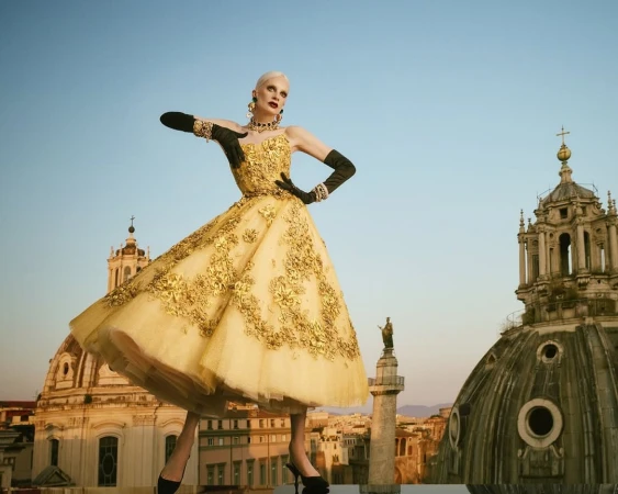 Kristen McMenamy in an ornate gold dress and black gloves on a rooftop with architectural domes under a clear sky.