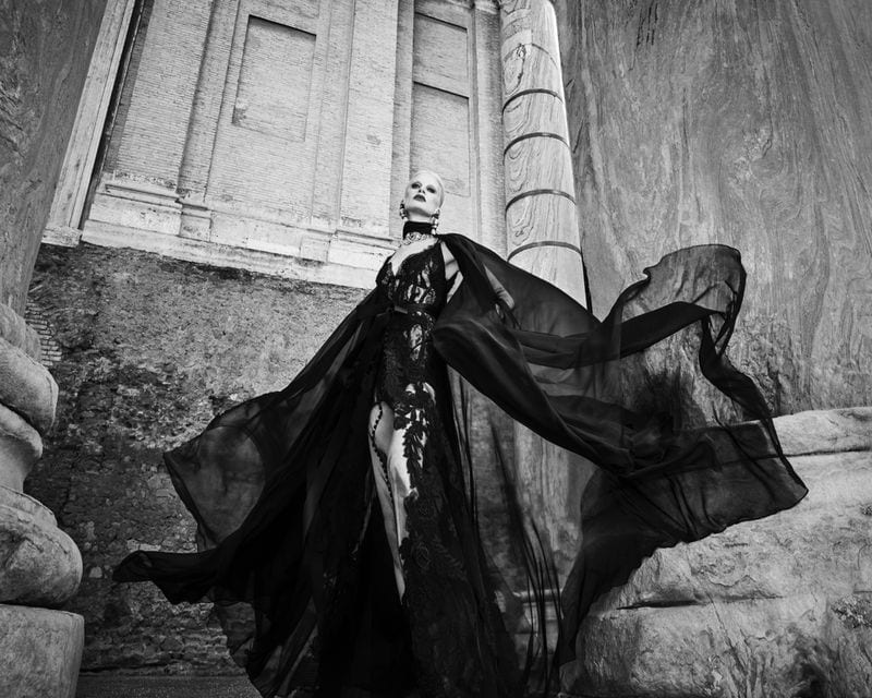 Monochrome fashion editorial image of Kristen McMenamy wearing a black gown with flowing cape standing amid classical stone architecture