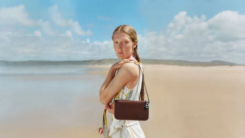 Model with a braided hairstyle wearing a white patterned dress holding a brown leather shoulder bag on a sandy beach