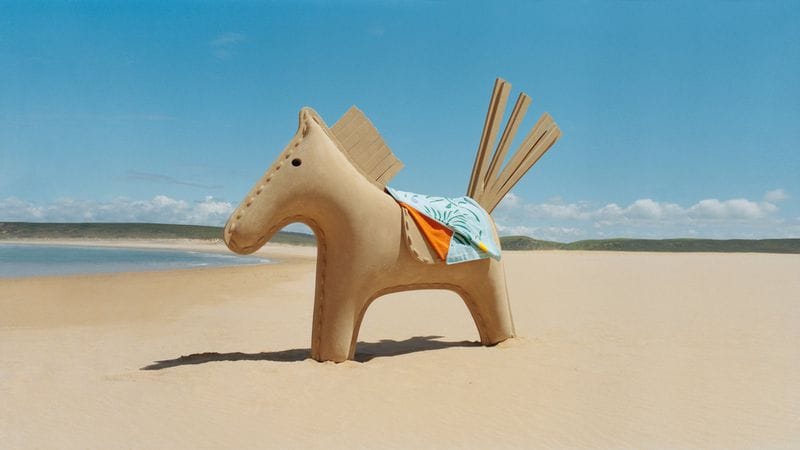 Sculptural sand horse sculpture standing on a sandy beach against an ocean and clear blue sky background.