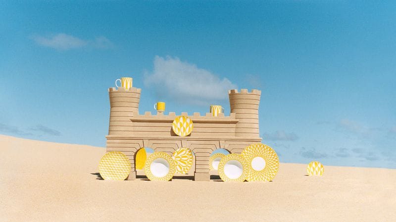 Sandcastle structure with yellow patterned plates and mugs set against sandy beach and clear blue sky.