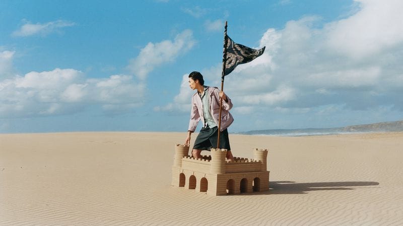 A man standing inside a large sandcastle holding a flag in a vast desert landscape under a blue sky.