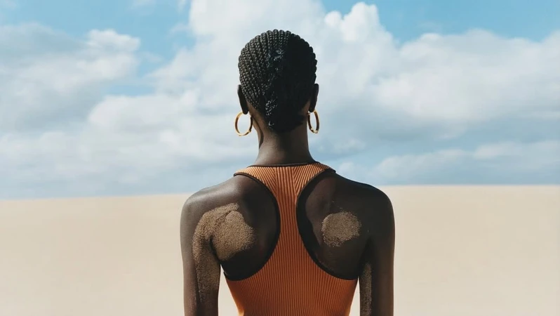 Model from behind wearing a brown ribbed tank top and gold hoop earrings against a sand dunes landscape and bright sky
