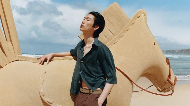 A man leaning against a large sand sculpture of a horse on a sunny beach