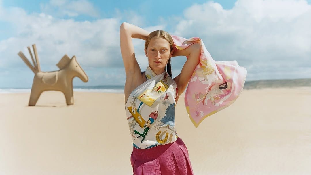 Model wearing a printed silk scarf top and pleated skirt on a sandy beach with a sculptural horse prop under an overcast sky.