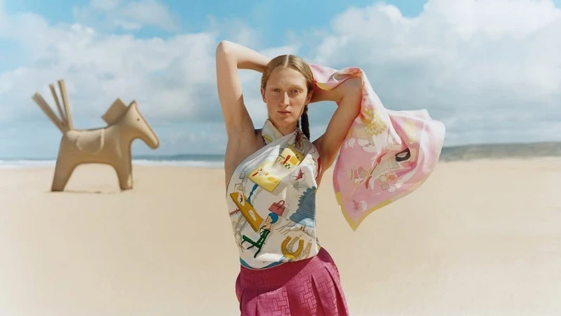 Model wearing a printed silk scarf top and pleated skirt on a sandy beach with a sculptural horse prop under an overcast sky.