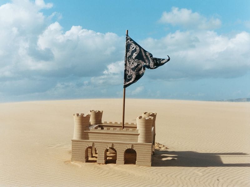 A detailed sandcastle with a dark patterned flag flying on a pole sits amidst vast sand dunes under a blue sky with white clouds.