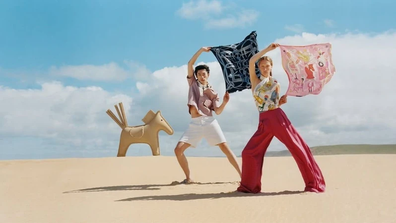 Two models holding up silk scarves against a sandy beach and a clear blue sky, with a sculptural horse prop.