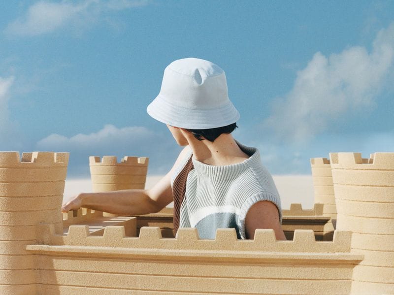 Model wearing a white bucket hat and knit vest inside a large sandcastle against a blue sky with clouds.