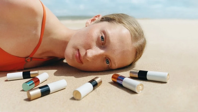 Model lying on sandy beach next to several lipstick tubes for Hermes Summer 2025 Ad Campaign by David Luraschi.